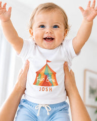 A smiling baby wearing a white t-shirt with a colorful circus tent graphic and the name " Josh" printed on it, being held up by someone's arms.