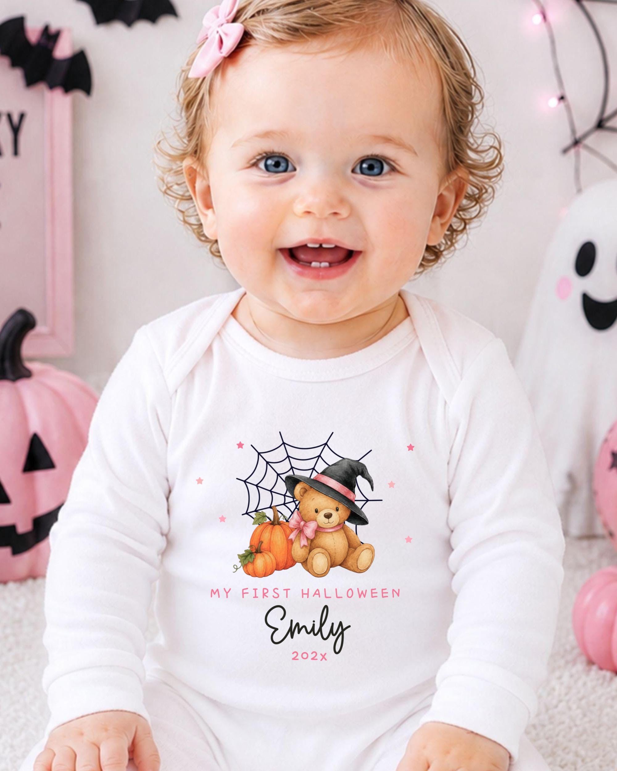 A smiling baby girl wearing a white onesie with a Halloween-themed design, including a teddy bear and pumpkins.