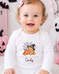 A smiling baby girl wearing a white onesie with a Halloween-themed design, including a teddy bear and pumpkins.