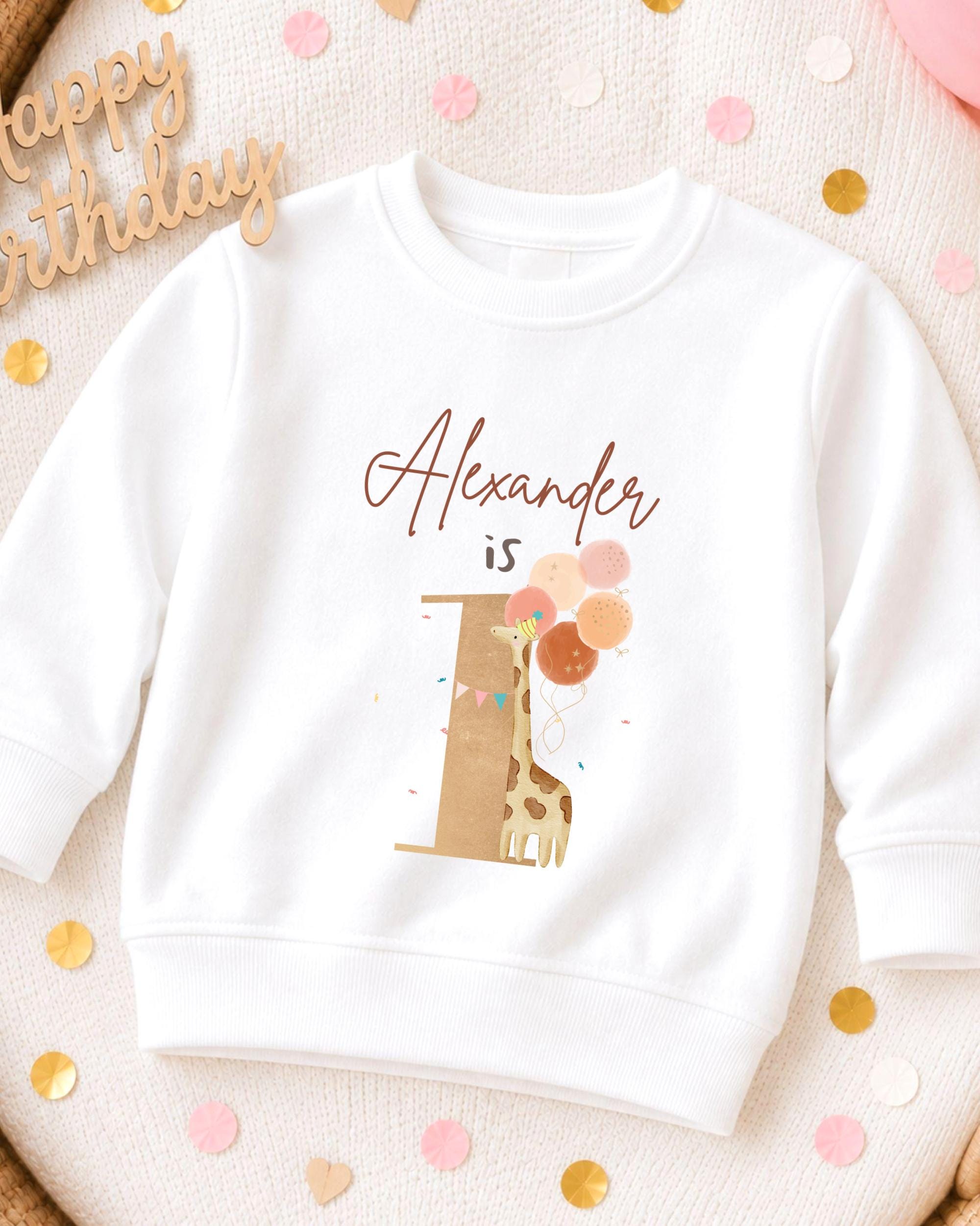 A white sweatshirt with a giraffe and balloons printed on it, along with the text "Alexander is" printed above the giraffe.
