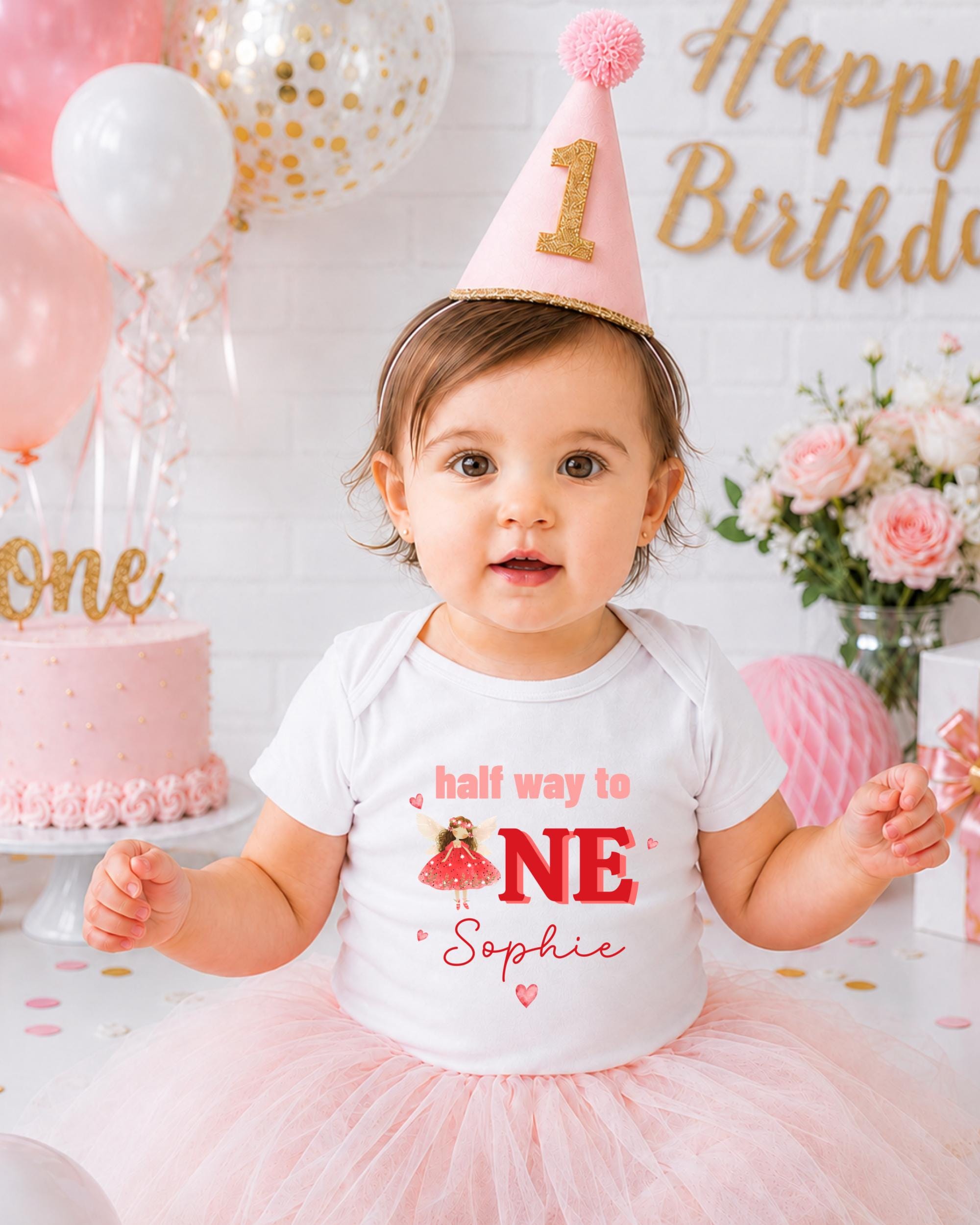 A young girl wearing a birthday hat and a white t-shirt with the text "half way to one" and "Sophie" is posing in front of a birthday cake and balloons.