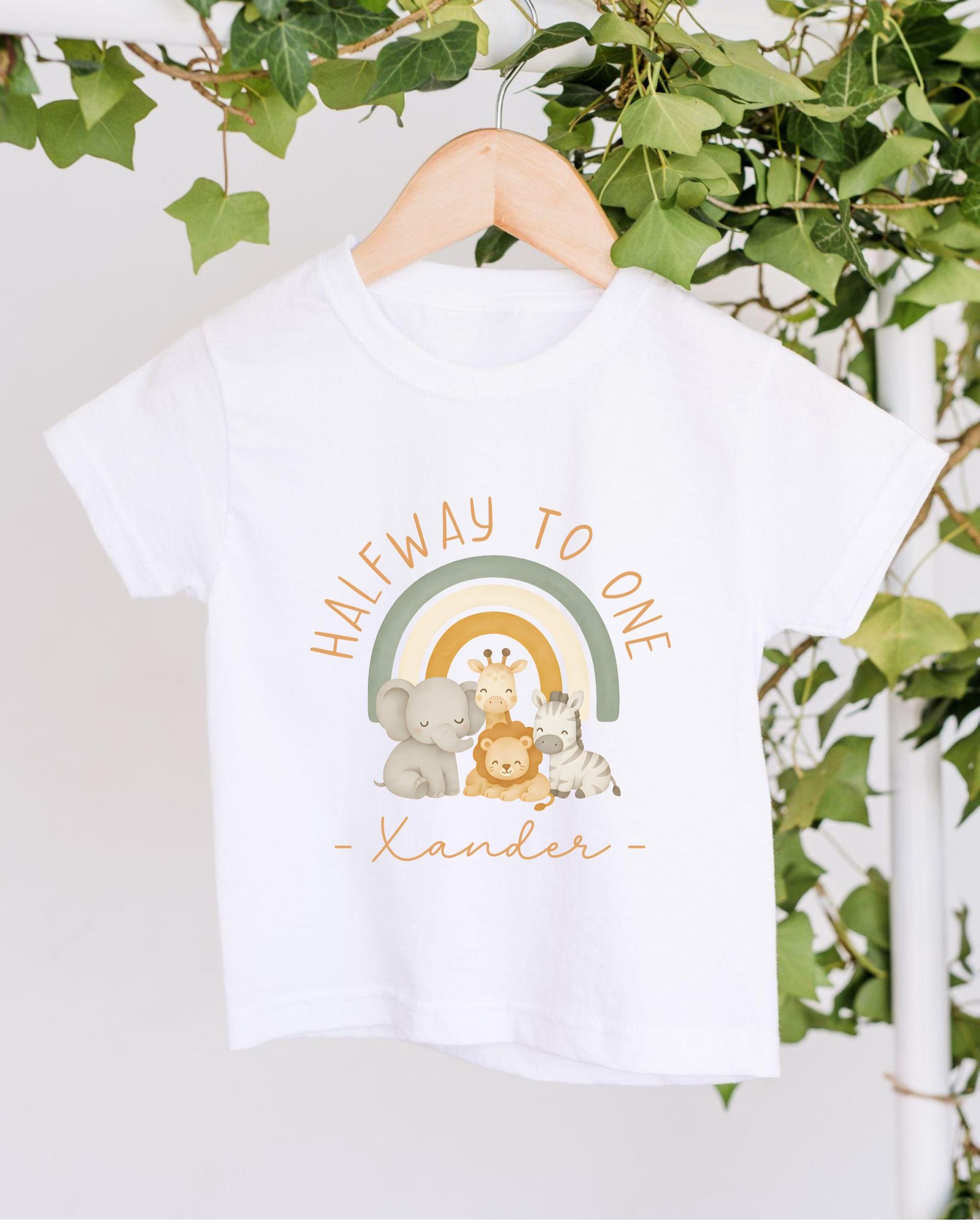 A white t-shirt with a graphic design featuring a rainbow and various animals, along with the text "Halfway to one" and the name "Xander" printed on it.