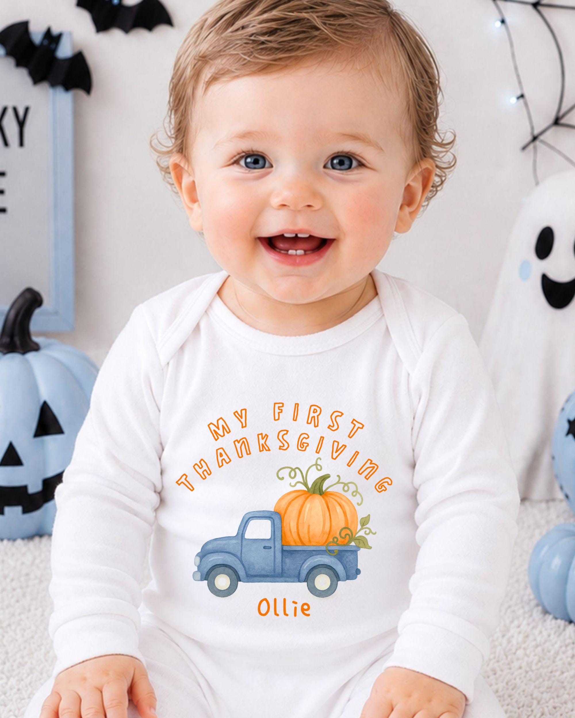 A smiling baby boy wearing a white onesie with a pumpkin and a truck graphic, sitting in front of Halloween decorations.
