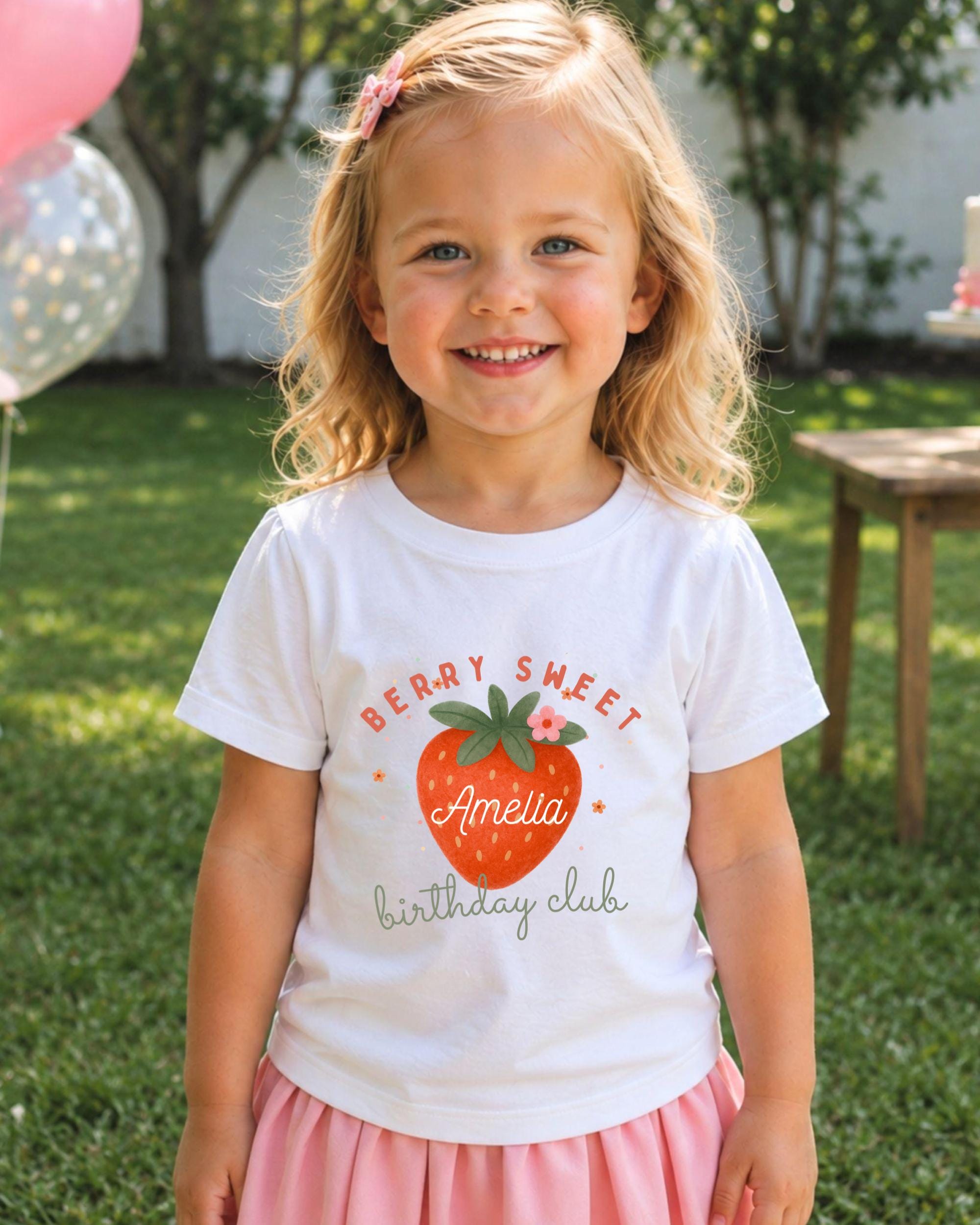 A young girl with blonde hair is smiling and wearing a white t-shirt with a strawberry graphic and text, standing in a grassy area with a wooden table and balloons in the background.