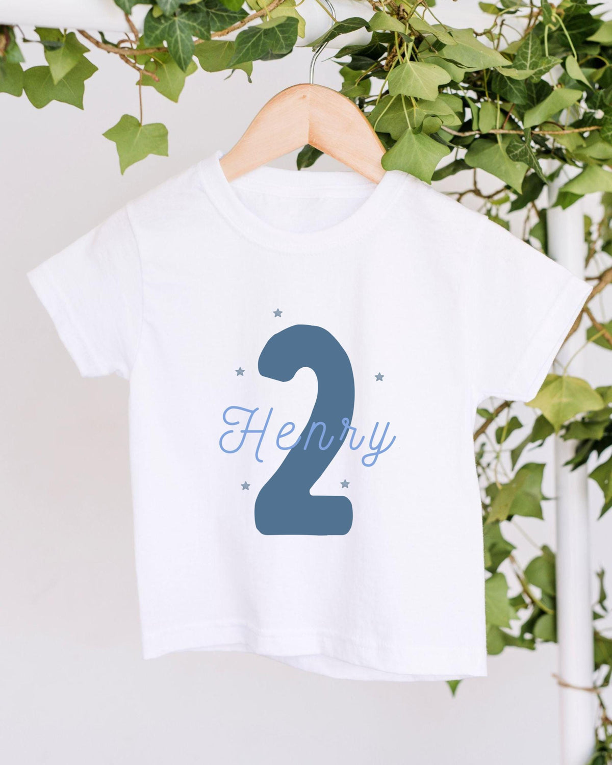 A white t-shirt with a blue number "2" and the name "HENRY" printed on it, hanging on a wooden hanger.