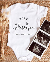 A white baby onesie with the text "BABY Harrison Due Sept 2027" printed on it, along with a photo of an ultrasound image.