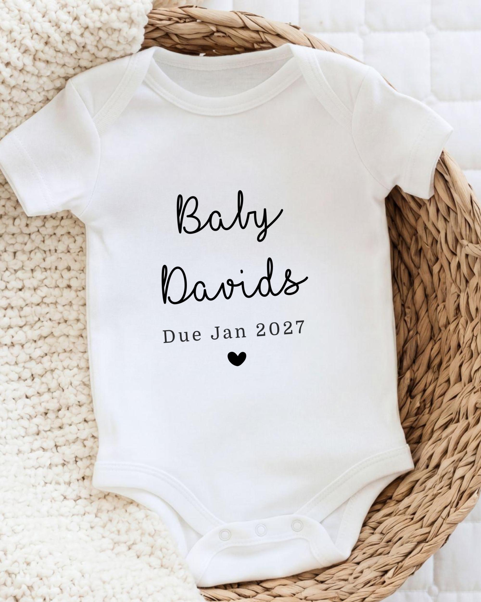 A white baby onesie with the text "Baby Davids Due Jan 2027" printed on it, placed on a woven basket.