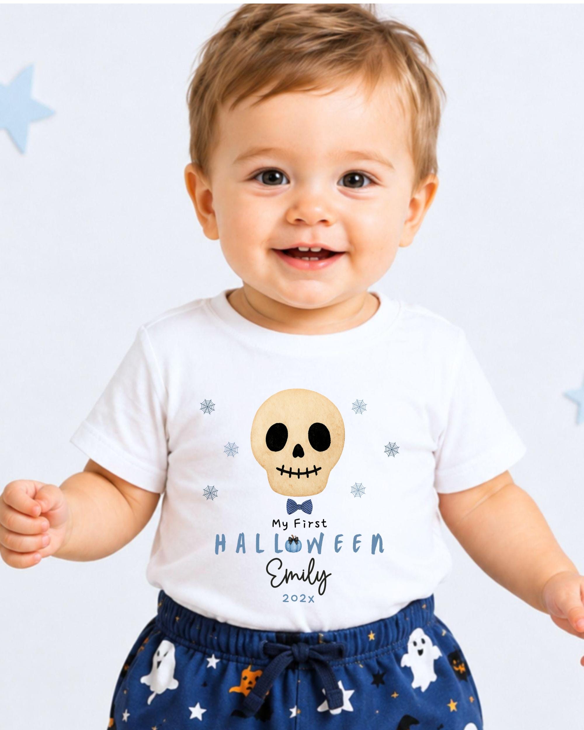 A smiling baby boy wearing a white t-shirt with a skull design and text, standing against a white background with blue stars.