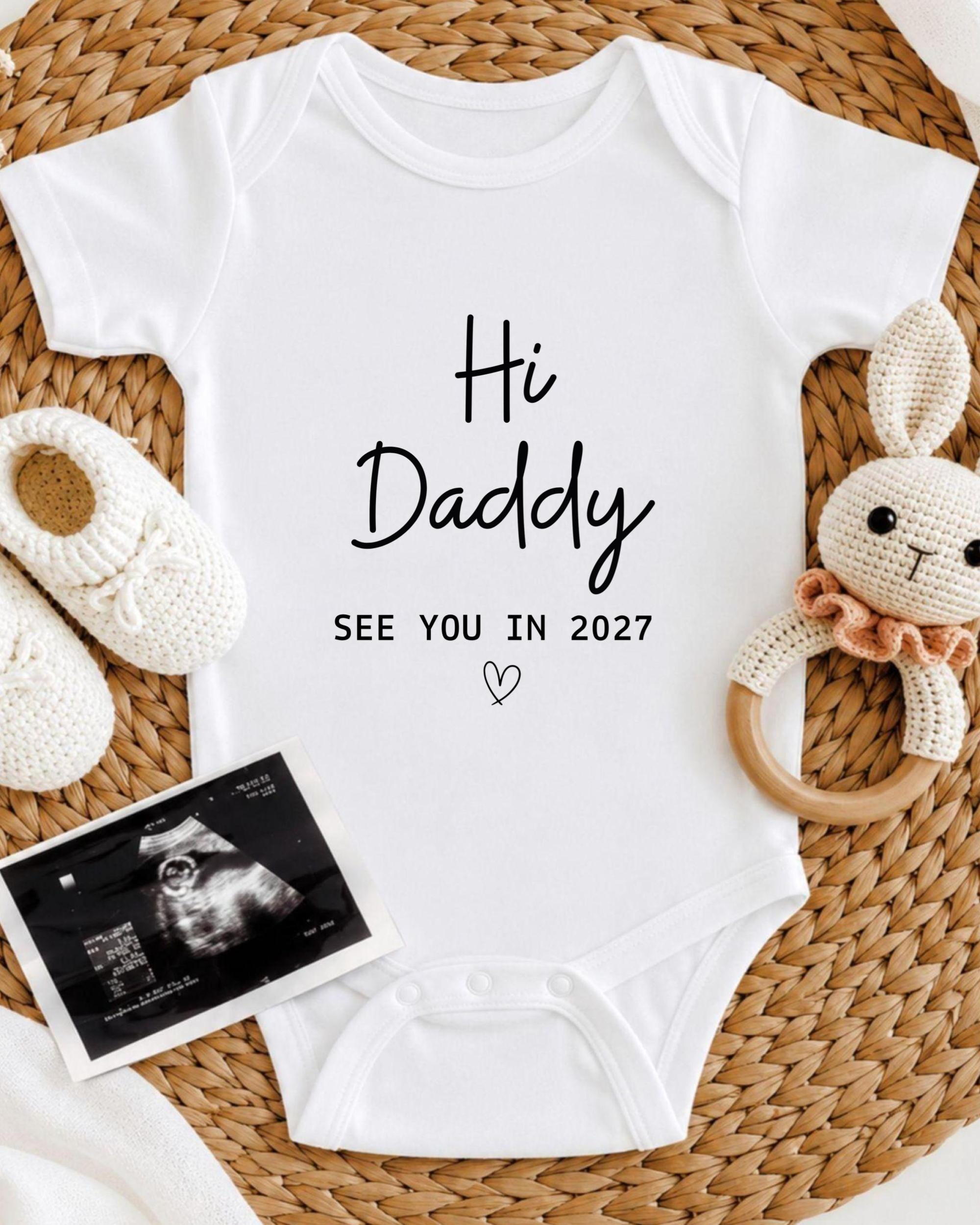 A white baby onesie with the text "Hi Daddy See You in 2027" printed on it, along with a crocheted bunny toy and a photograph of an Ultrasound image.