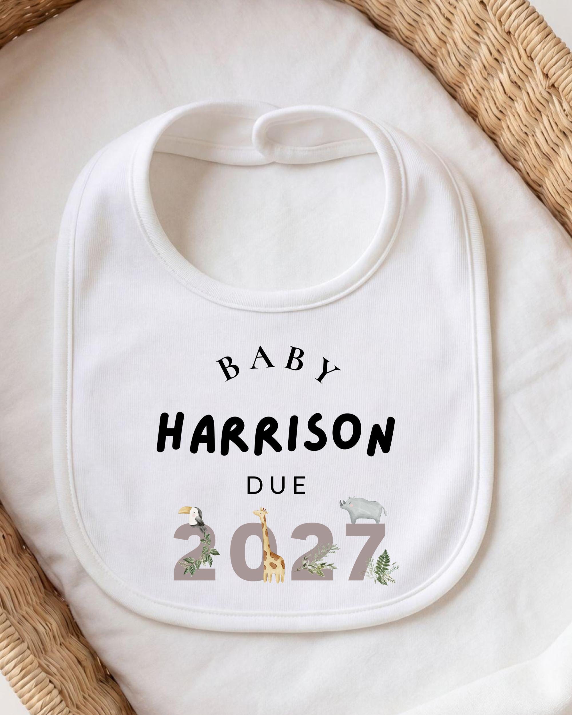 A white bib with the text "BABY HARRISON DUE 2027" printed on it, along with various animal illustrations.