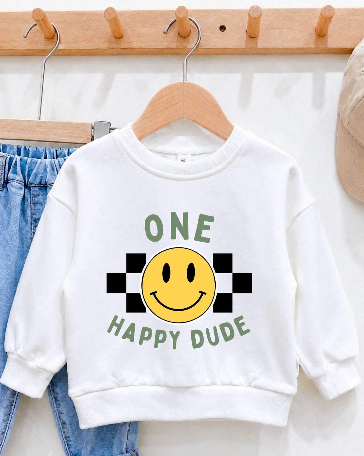 A white sweatshirt with a cheerful yellow smiley face and the text "ONE HAPPY DUDE" printed on it, hanging on a wooden hanger.