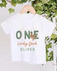 A white t-shirt with a graphic design featuring a cartoon duck wearing a party hat and the text "One Lucky Duck Oliver" printed on it.