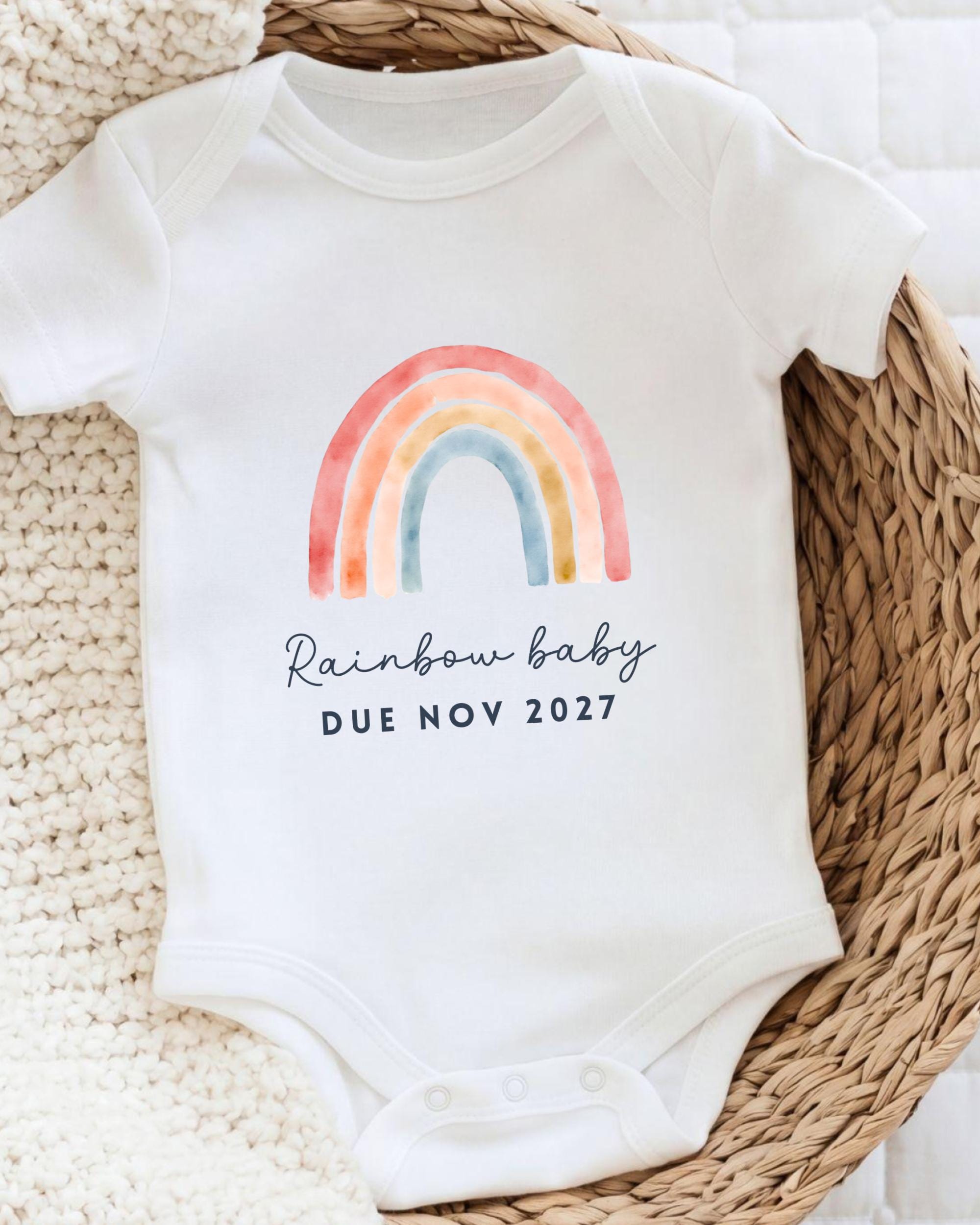 A white baby onesie with a colorful rainbow graphic and the text "Rainbow baby due novely 2027" printed on it.