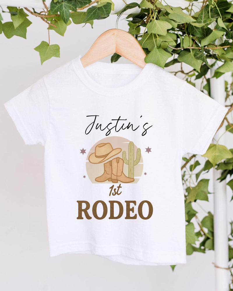 A white t-shirt with a graphic design featuring a cowboy hat, a cactus, and the text "Justin's 1st Rodeo" printed on it.