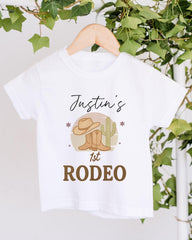 A white t-shirt with a graphic design featuring a cowboy hat, a cactus, and the text "Justin's 1st Rodeo" printed on it.
