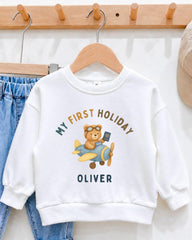 A white sweatshirt with a graphic of a teddy bear wearing goggles and a pilot's hat, along with the text "My first holiday Oliver" printed on it.