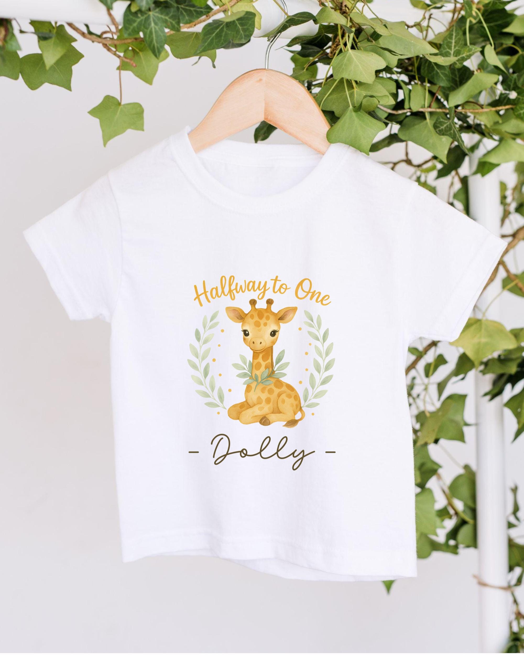 A white t-shirt with a giraffe graphic and the text "Halfway to One - Dolly" printed on it, hanging on a wooden hanger.