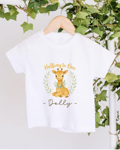 A white t-shirt with a giraffe graphic and the text "Halfway to One - Dolly" printed on it, hanging on a wooden hanger.