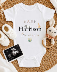 A baby announcement t-shirt with the name "Baby Harrison" and an image of a baby surrounded by baby-related items, including a crocheted bunny, white baby booties, and an Ultrasound image.
