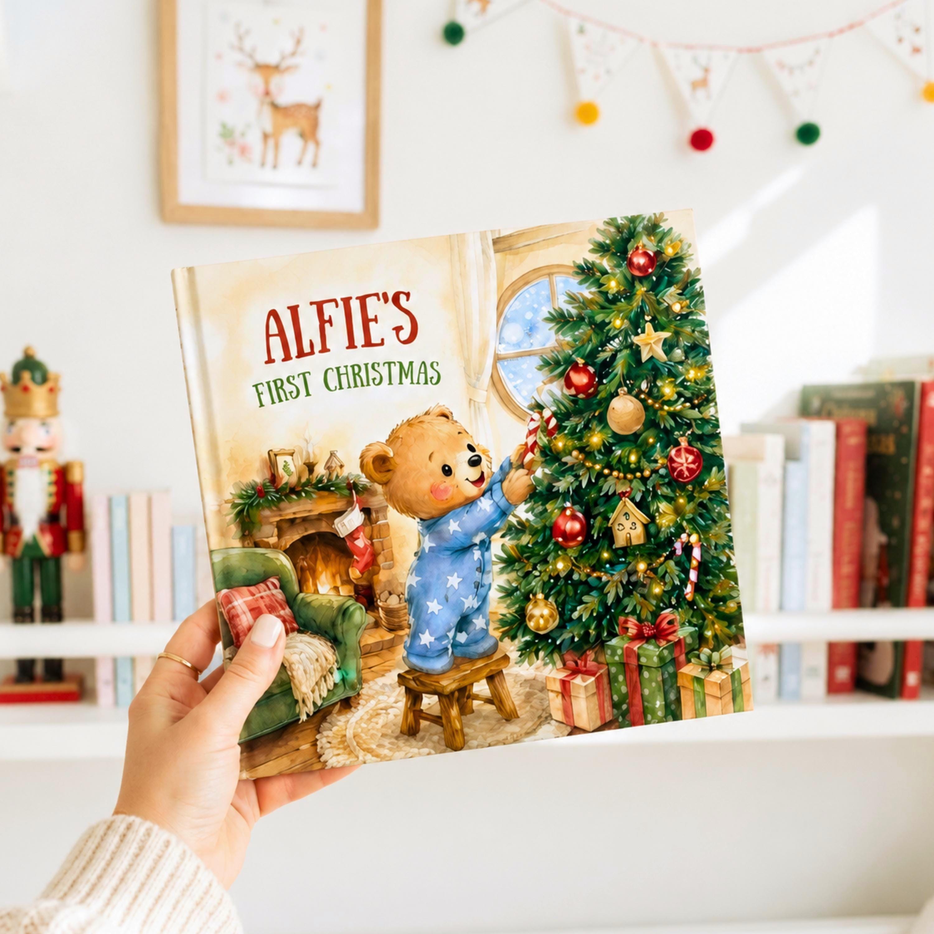 A hand holding a book titled "Alfie's First Christmas" with an illustration of a teddy bear decorating a Christmas tree.