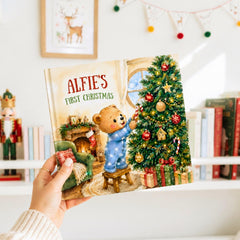 A hand holding a book titled "Alfie's First Christmas" with an illustration of a teddy bear decorating a Christmas tree.