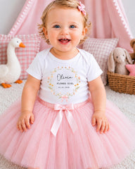 A smiling baby girl wearing a white t-shirt with a floral design and a pink tulle skirt, sitting on a pink carpet surrounded by stuffed animals.
