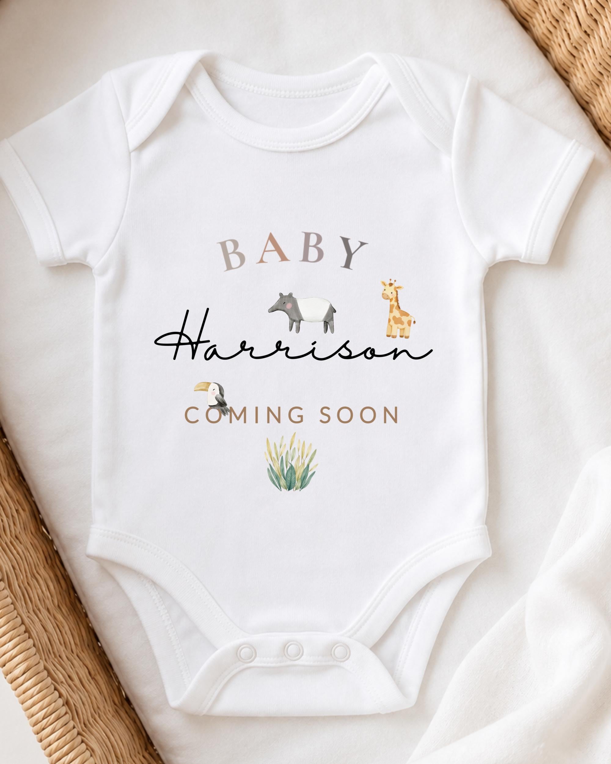 A white baby onesie with the text "BABY Harrison" and illustrations of a giraffe and a bird, along with the text "Coming Soon" printed on it.