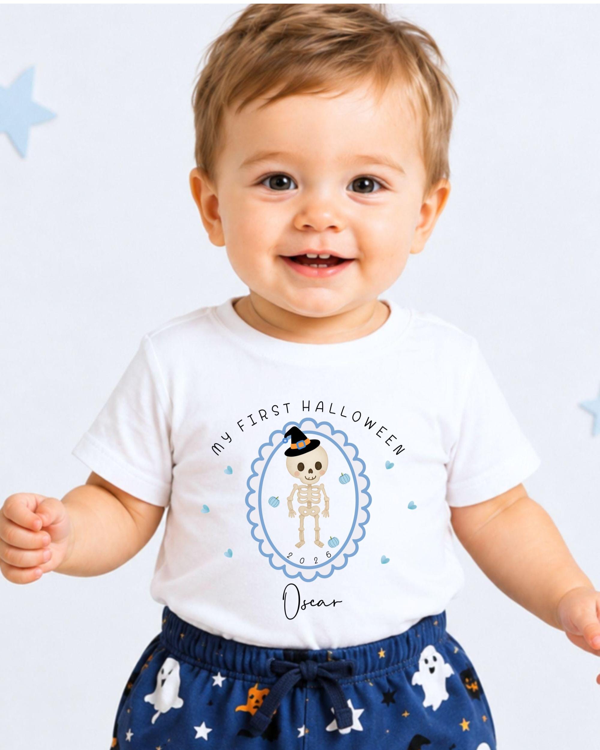 A smiling baby boy wearing a white t-shirt with a Halloween-themed graphic and text, standing against a white background with blue stars.