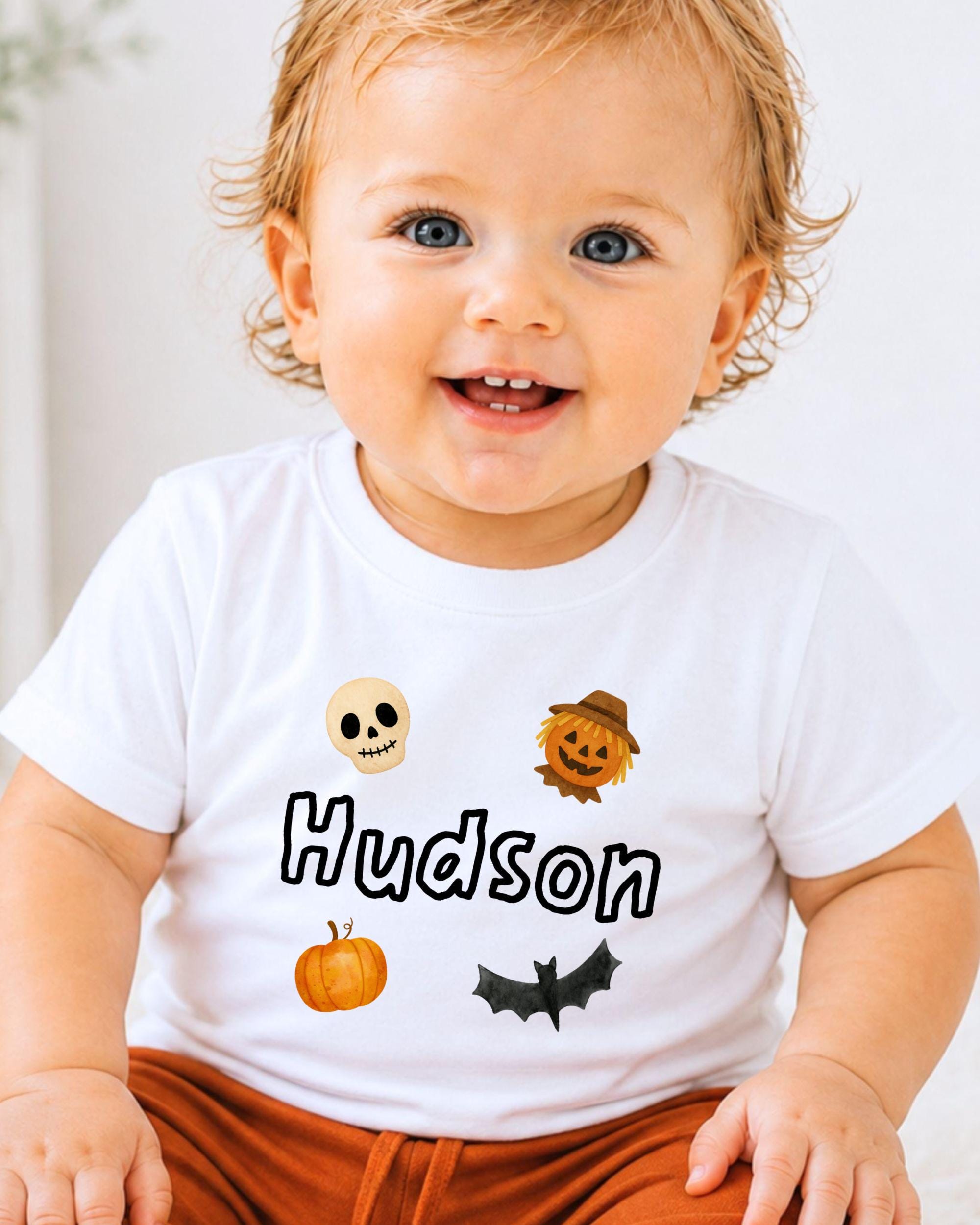 A smiling baby boy wearing a white t-shirt with Halloween-themed icons and the name "Hudson" printed on it.