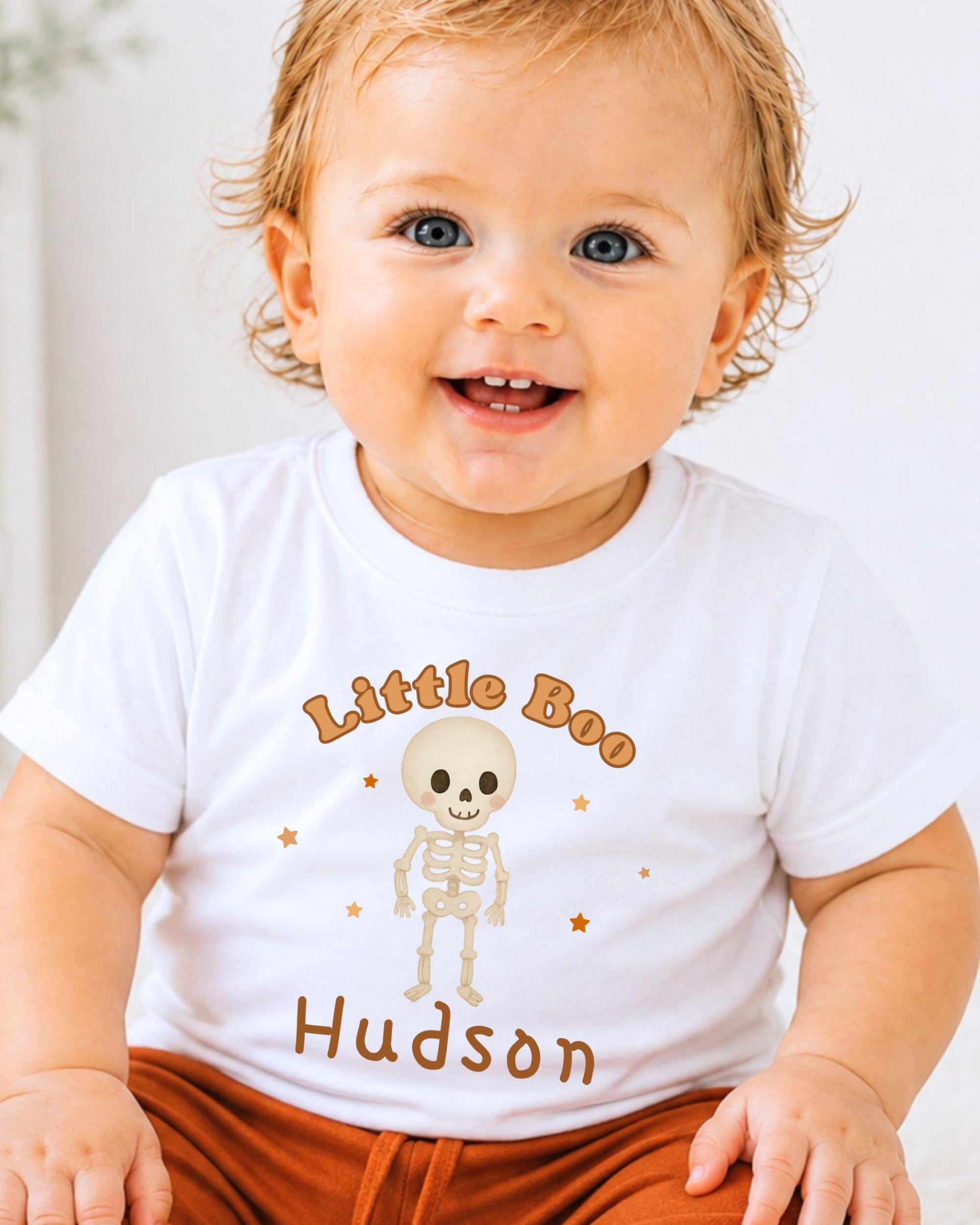 A smiling baby wearing a white t-shirt with a skeleton graphic and the name "Hudson" printed on it.