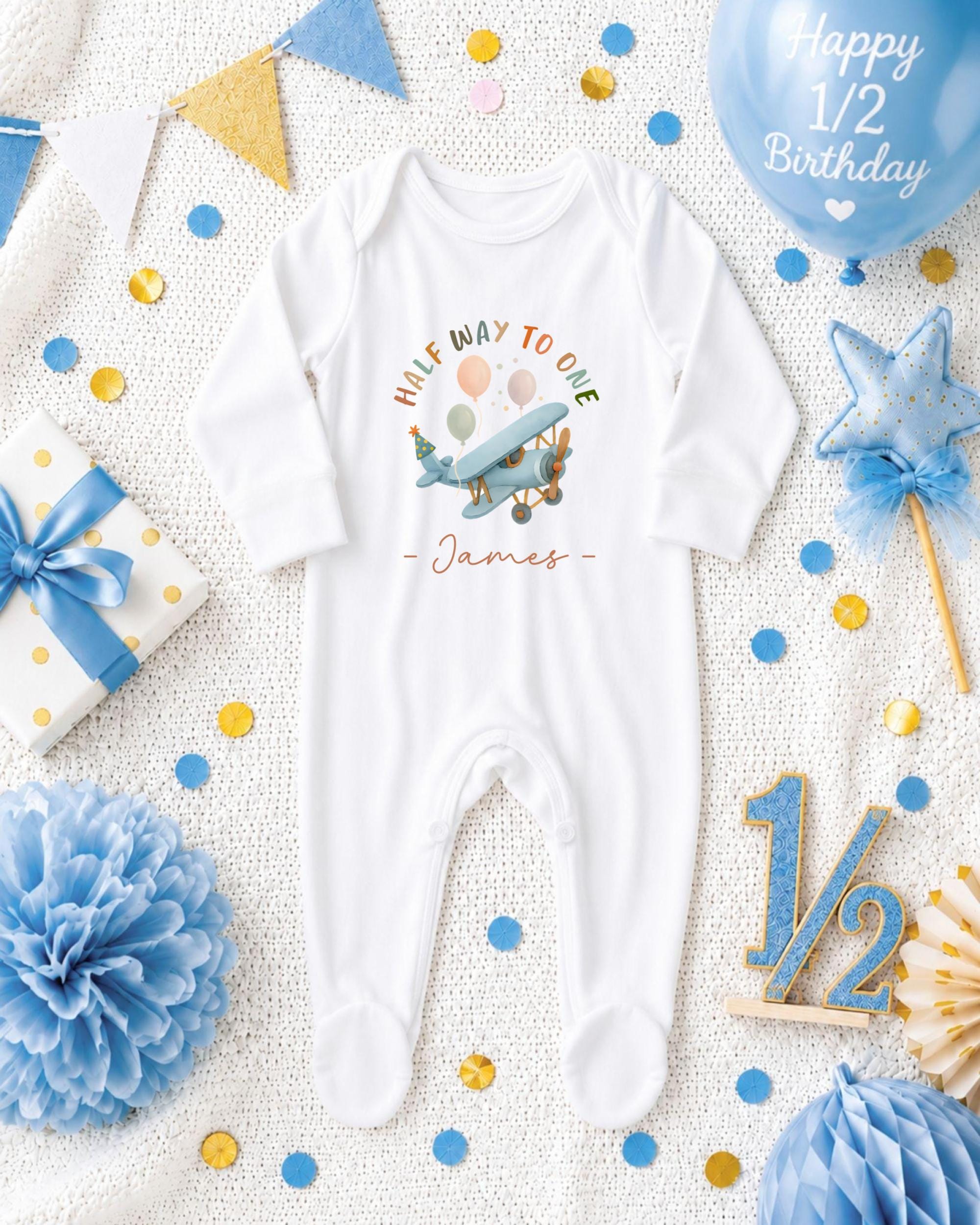 A baby onesie with a half-way to one airplane design, surrounded by confetti, a blue balloon, and other baby-themed items.
