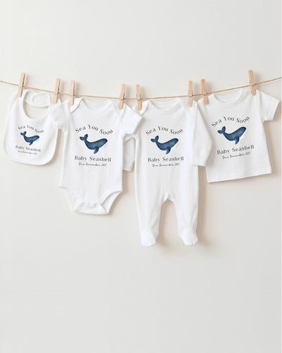 Personalisable white Bib, Baby Vest, Baby Grow and T Shirt with a blue watercolour whale between black 'Sea You Soon' and 'Baby Seashell Due December 2027' lettering.
