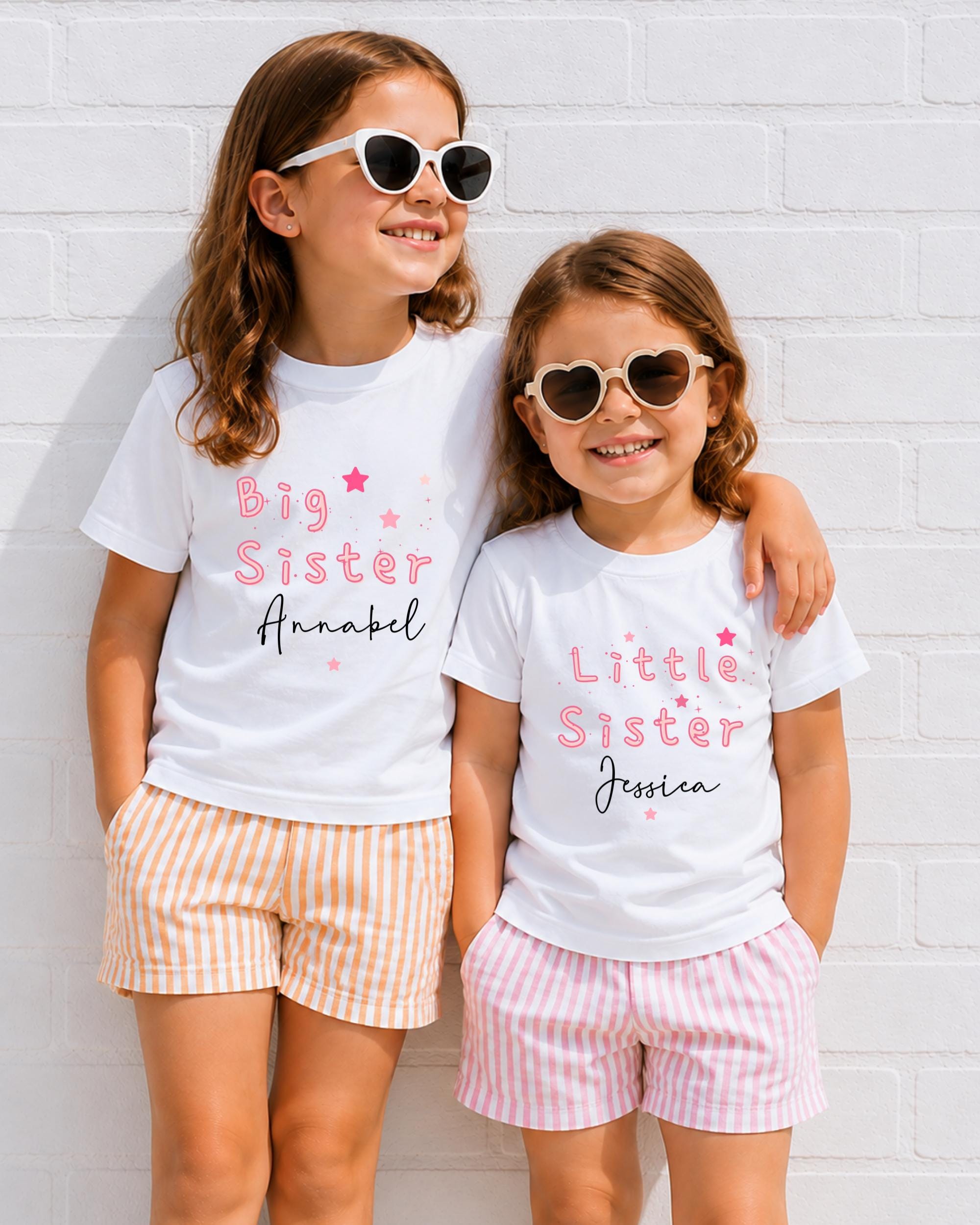Two young girls wearing matching white t-shirts with the text "Big Sister Annabel" and "Little Sister Jessica" printed on them, smiling and posing together against a white brick wall.