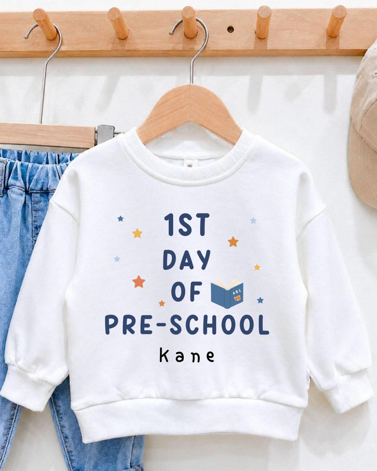 A white sweatshirt with the text "1st DAY OF PRE-SCHOOL kane" printed on it, along with stars and a book icon.