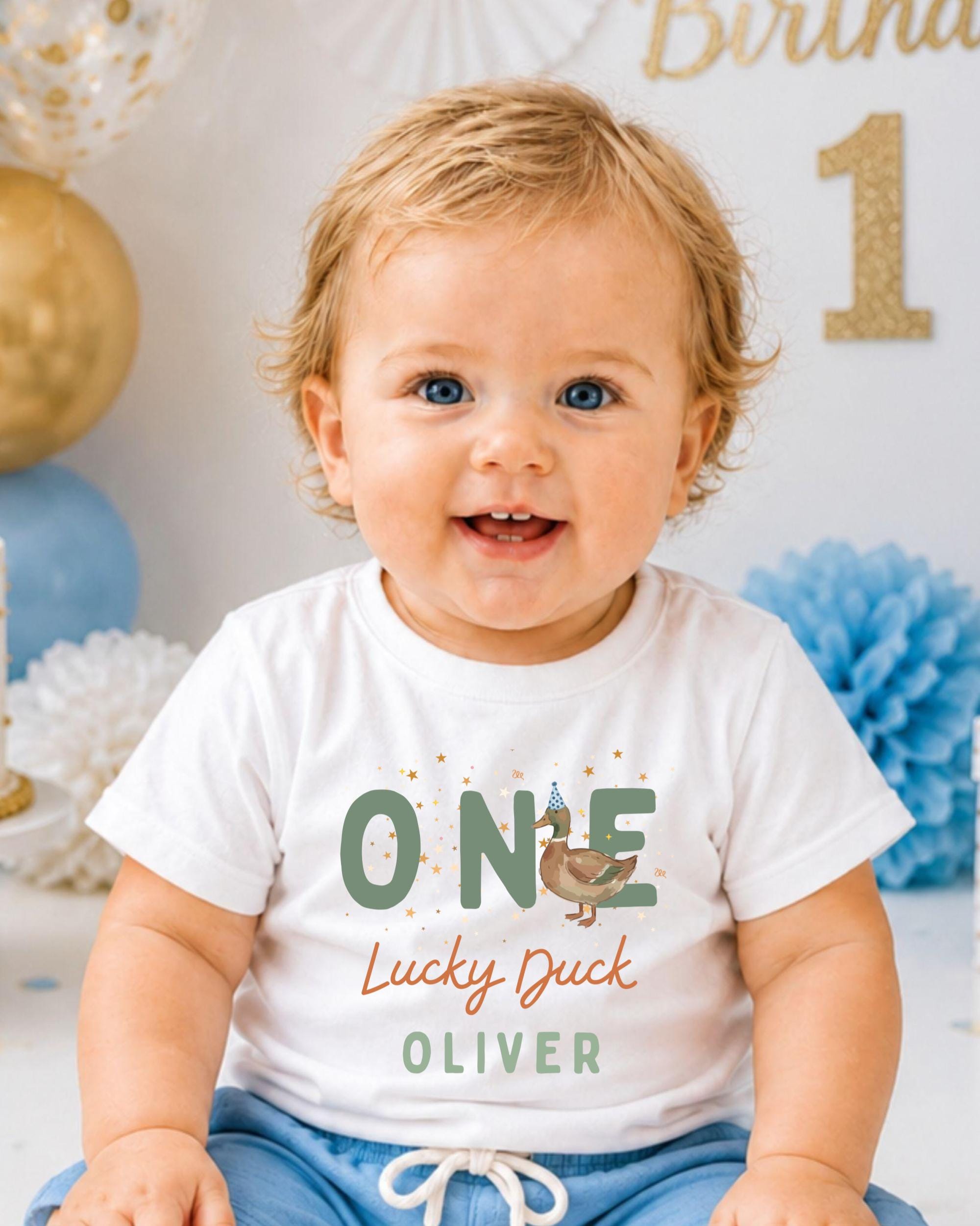A smiling baby boy wearing a white t-shirt with a graphic of a duck and the text "One Lucky Duck Oliver" is sitting in front of a birthday party backdrop.