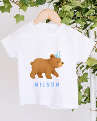 A white t-shirt with a cartoon bear wearing a party hat and the name "WILSON" printed on it, hanging on a wooden hanger.