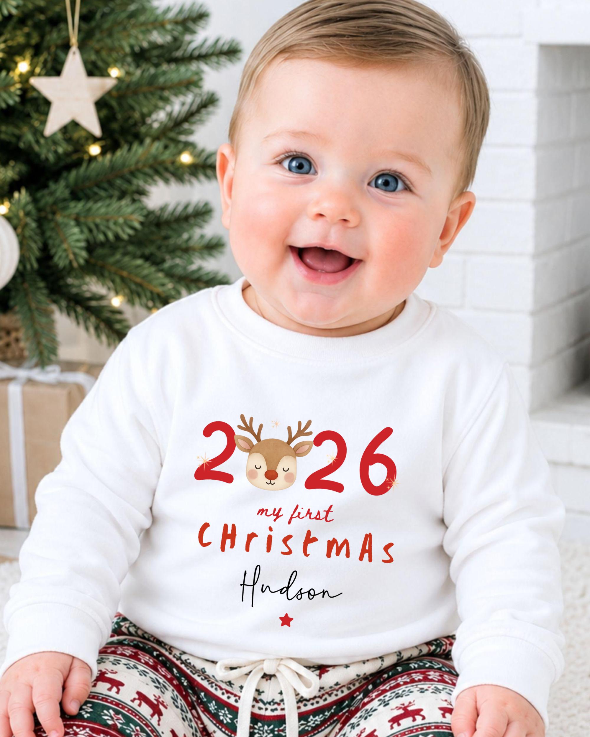 A smiling baby boy wearing a white sweater with the text "26 my first christmas Hudson" printed on it, sitting in front of a Christmas tree.
