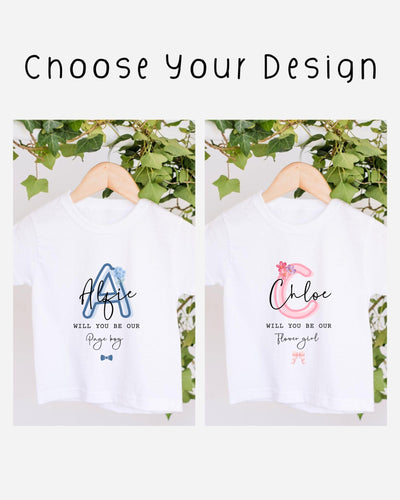 two white t-shirts hanging on a wooden hanger, each with a personalized message printed on them. The messages read "Will you be our Page boy" and "Will you be our Flower girl", suggesting that the t-shirts are intended as gifts or souvenirs for someone named Page boy and Flower girl.