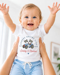 A smiling baby wearing a white t-shirt with a graphic of a race car and the text "Sunday Racing Club EST. Daddy & Hudson" being held up by two people.