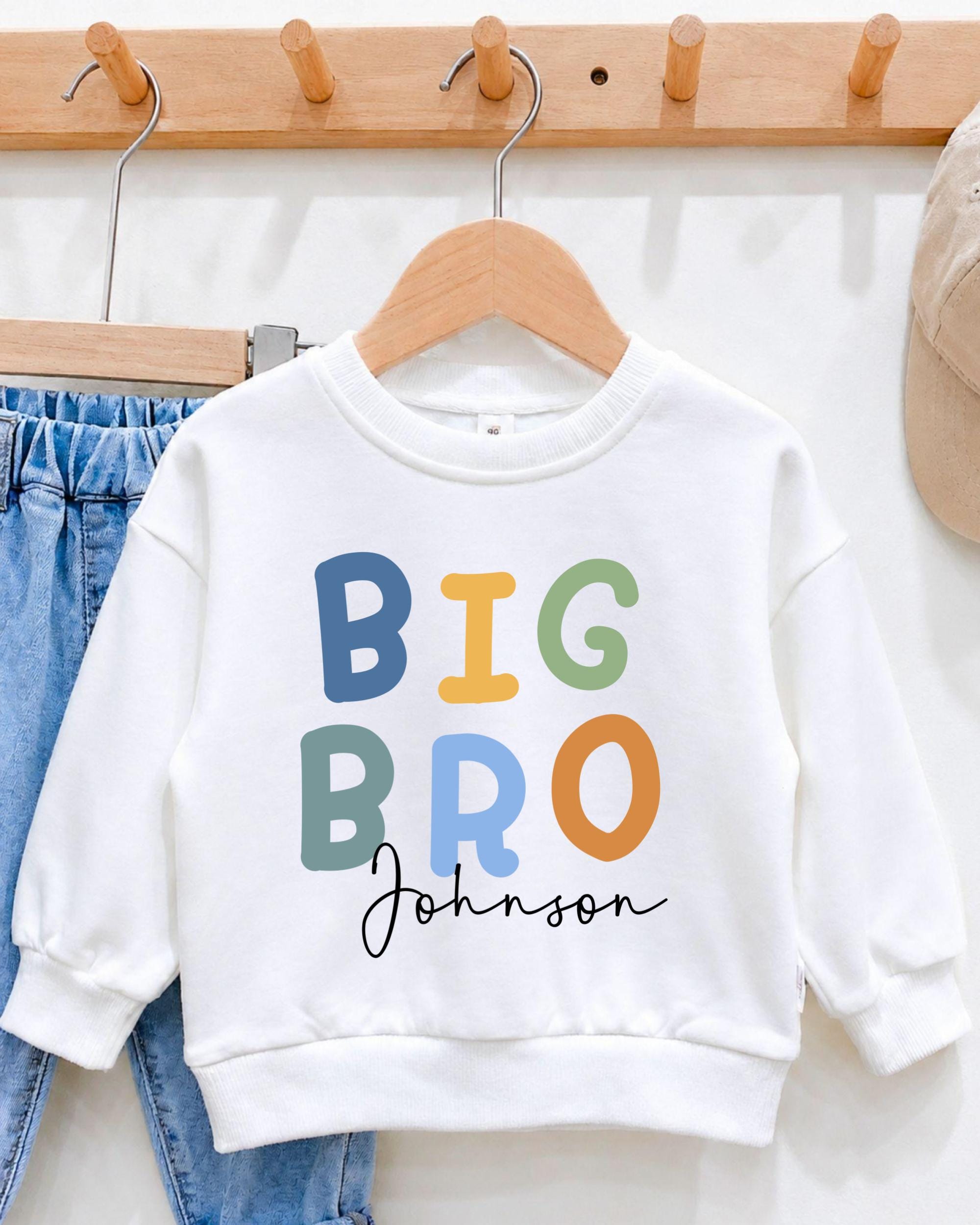 A white sweatshirt with the text "BIG BRO Johnson" printed on it, hanging on a wooden hanger.