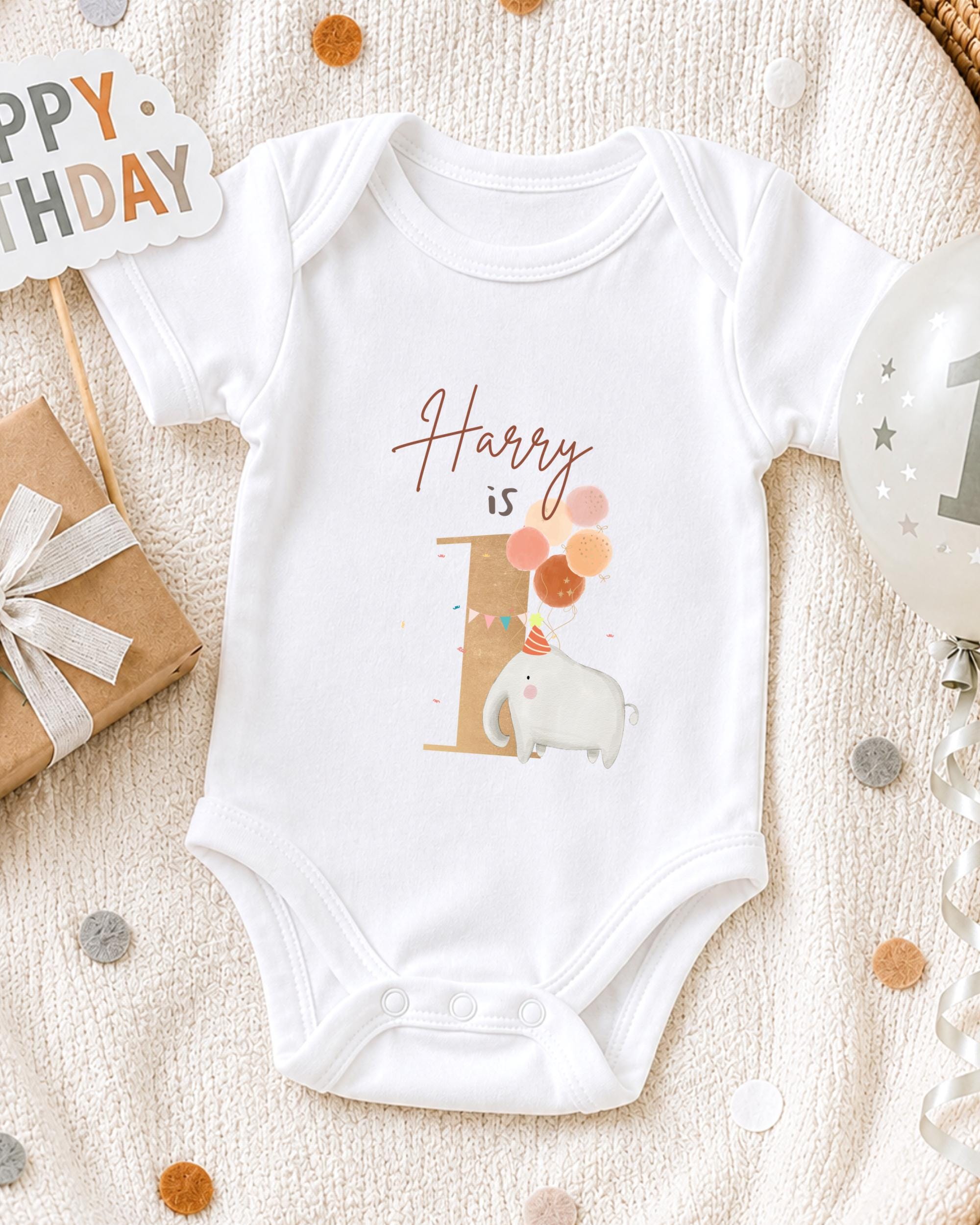 A white onesie with a personalized name and a cartoon elephant printed on it, along with a "Happy Birthday" sign and a birthday balloon.