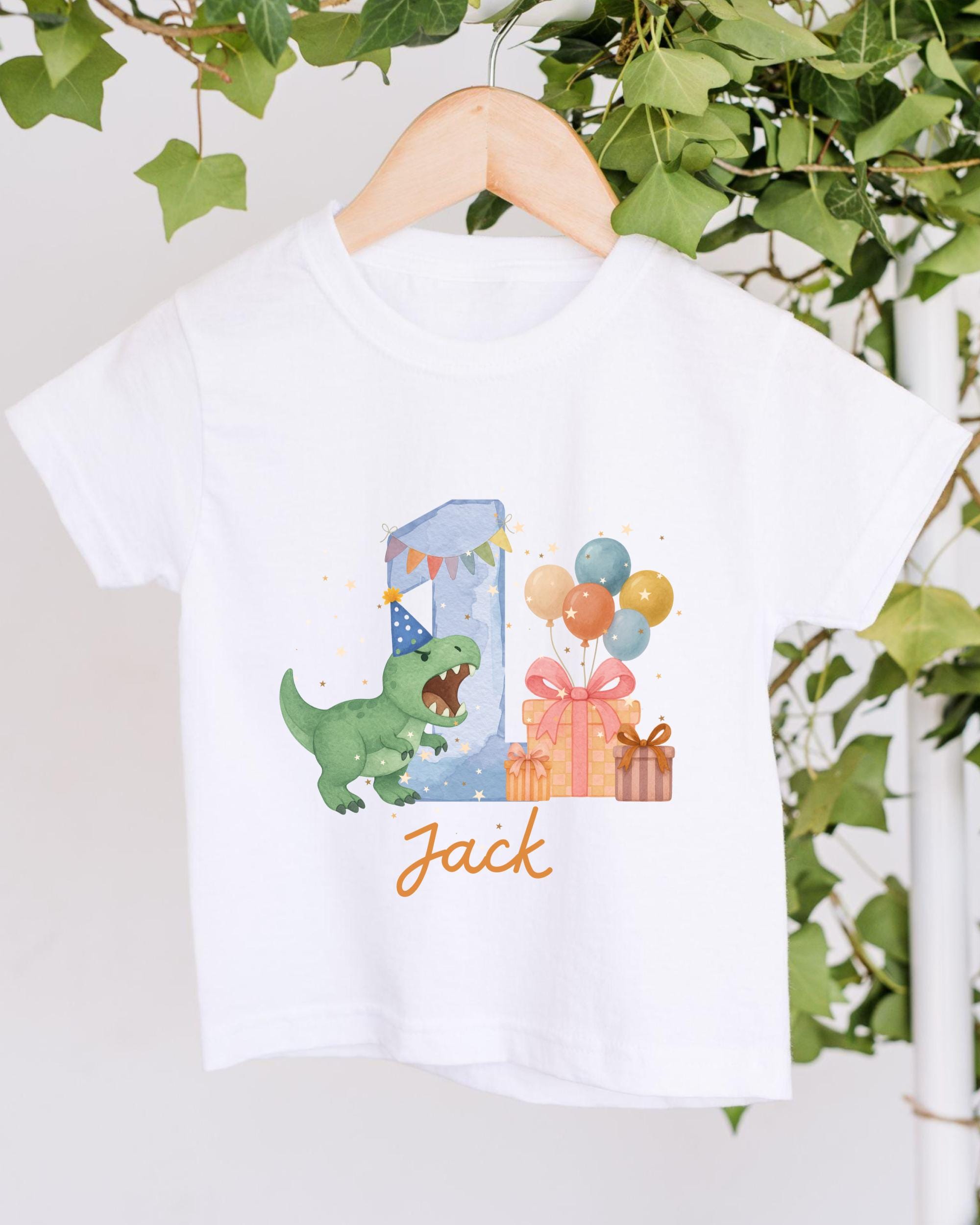 A white t-shirt with a graphic design depicting a dinosaur wearing a party hat and surrounded by colorful balloons and presents.