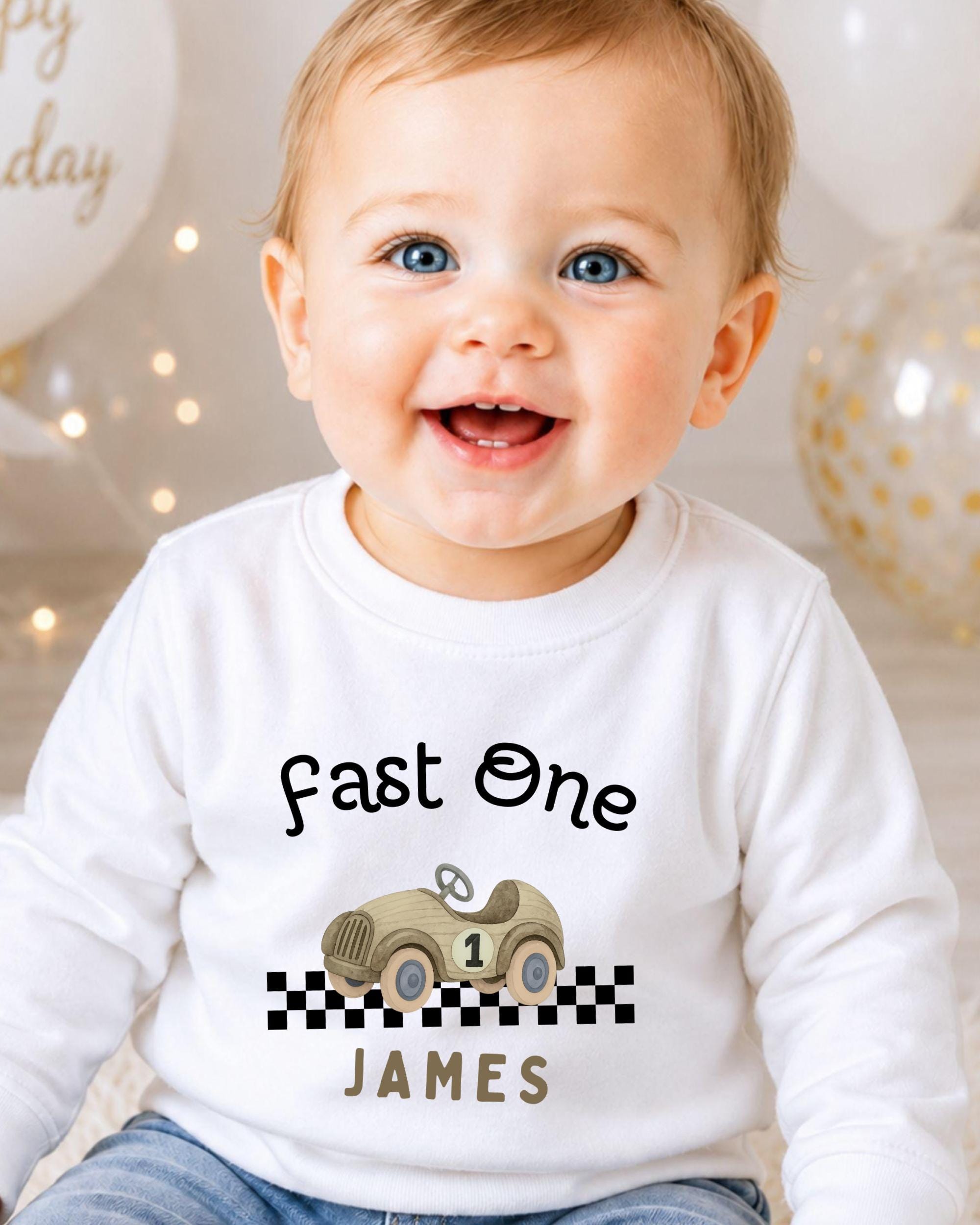 A smiling baby boy wearing a white shirt with a race car graphic and the text "Fast One James" printed on it.