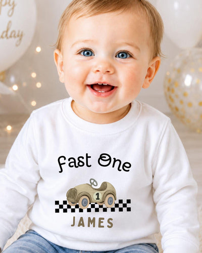A smiling baby boy wearing a white shirt with a race car graphic and the text "Fast One James" printed on it.