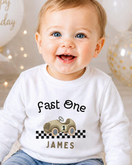 A smiling baby boy wearing a white shirt with a race car graphic and the text "Fast One James" printed on it.