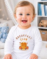 A smiling baby wearing a white onesie with a breakfast club graphic and text.