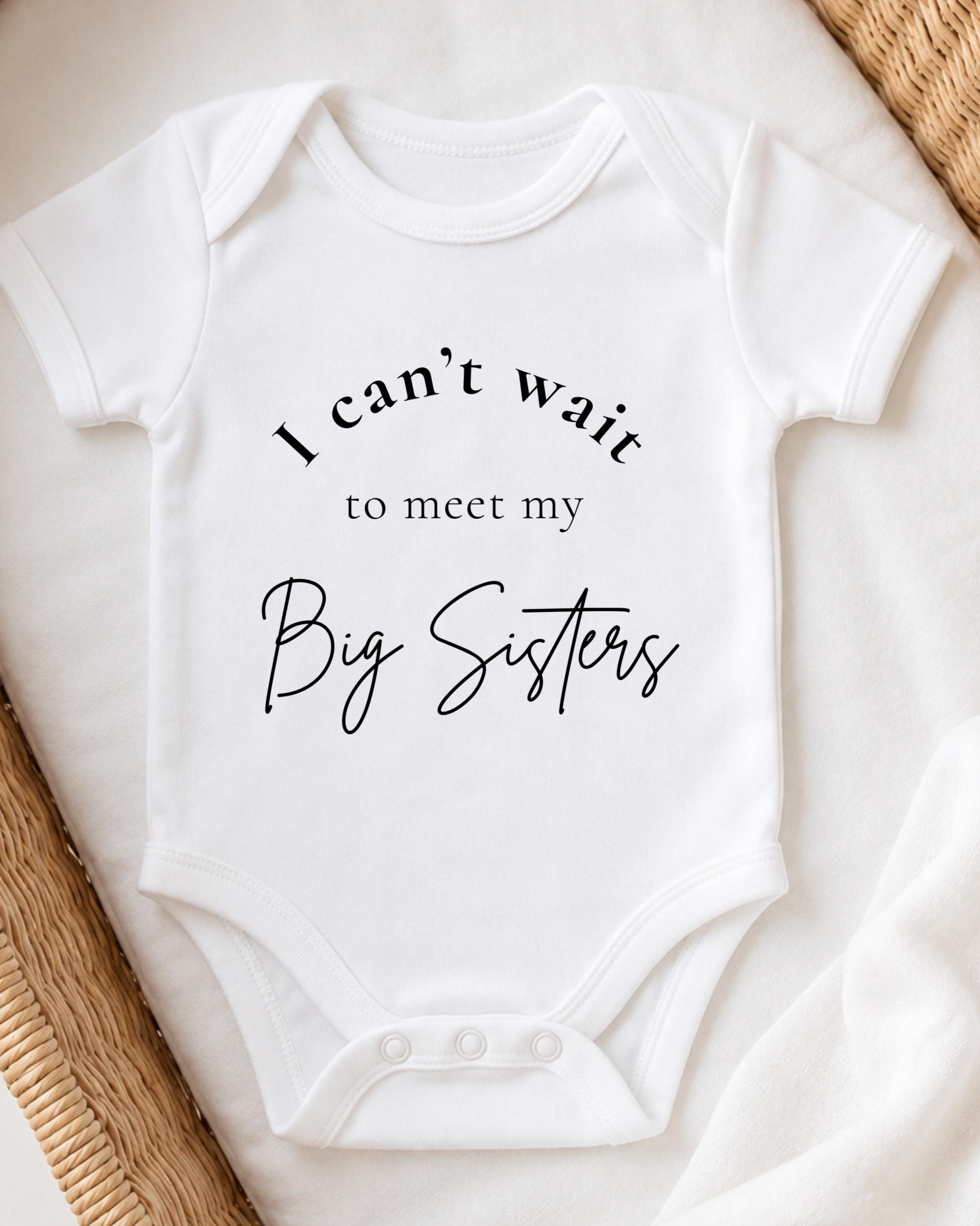 A white baby onesie with the text "I can't wait to meet my Big Sisters" printed on it.
