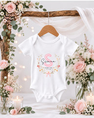 A white baby onesie with a pink floral design hangs on a wooden hanger, surrounded by pink and white flowers, candles, and greenery.