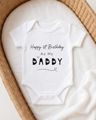 A white onesie with the text "Happy 1st Birthday As My Daddy" printed on it, placed in a woven basket.