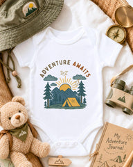 a baby onesie with a camping scene printed on it, surrounded by various camping-related items such as a hat, compass, binoculars, and a teddy bear.